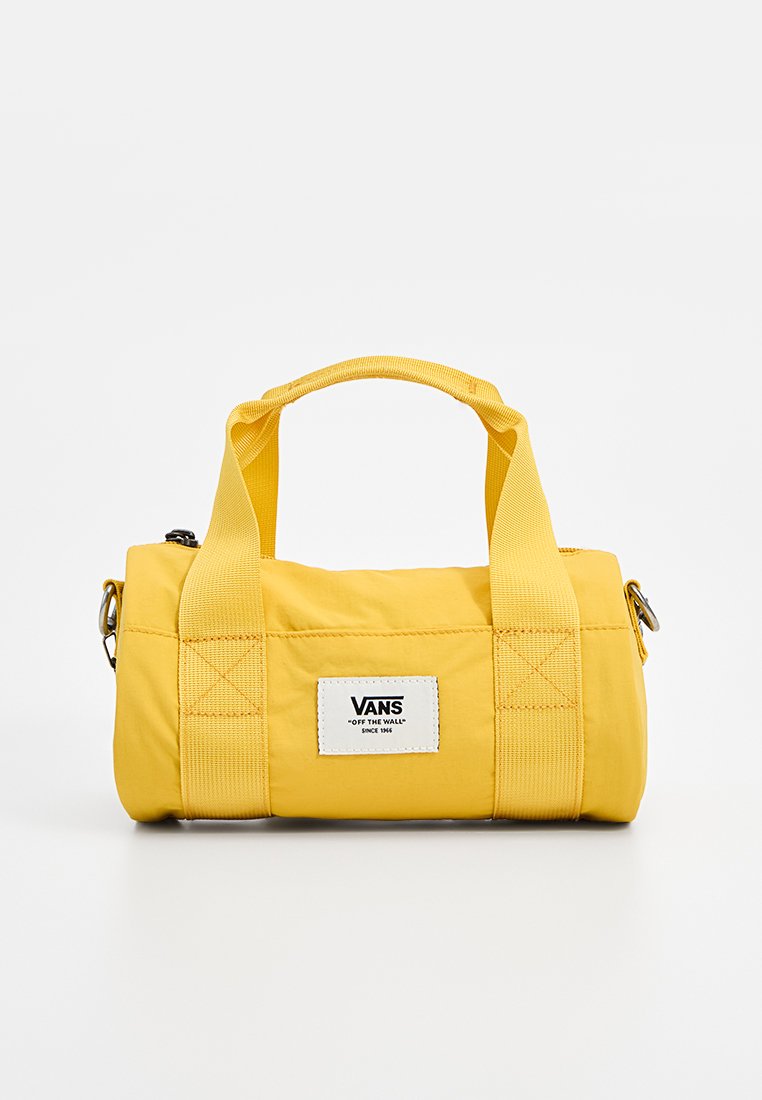 Gul tygresväska med två korta handtag, platt rektangulär botten och en vit etikett med "VANS." Slen yta, enkelt design.