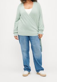 Mintgrön omslagströja i mjukt, texturerat stickat, parat med ljusblå influenser jeans i lös passform och vita sneakers med beige sulor.