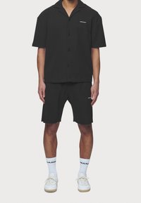 Czarna koszula na krótki rękaw z kołnierzykiem i guzikami, zestawiona z czarnymi szortami. Obie części wykonane są z fakturowanej tkaniny i mają drobne logo. Białe trampki i skarpetki.