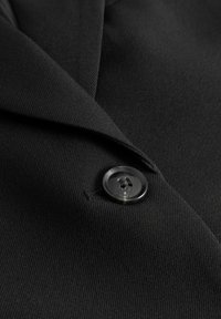 Primer plano de la solapa de una chaqueta de traje negra con un botón negro, brillante, de dos agujeros, abrochado.