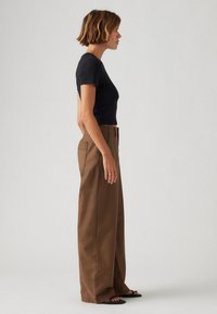 Schwarzes, tailliertes T-Shirt kombiniert mit hochgeschnittenen, braunen, weitem Hosen, die über eine glatte Textur und Fronttaschen verfügen. Einfaches Design, minimale Akzente.