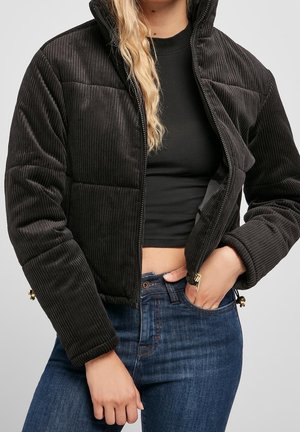 Ung person iført en sort cropped fløjlsjakke over en sort cropped top og blå jeans, med den ene hånd i lommen.