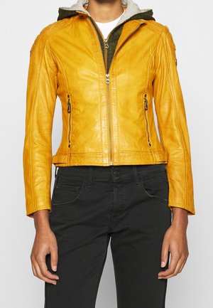 Persona vistiendo una chaqueta de cuero amarilla sobre una sudadera con capucha verde y pantalones negros, de pie contra un fondo liso y claro.