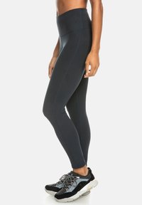 Hoch taillierte schwarze Leggings aus dehnbarem Material, mit Nahtdetails und glatter Textur. Kombiniert mit schwarzen und silbernen Sportschuhen.