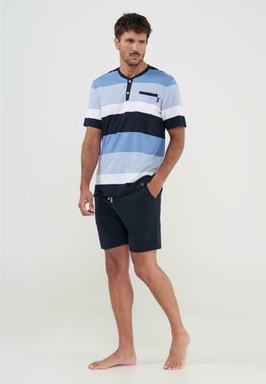 Hombre de pie descalzo, vistiendo una camisa de manga corta a rayas azules, blancas y negras con pantalones cortos azul marino, una mano en el bolsillo, fondo neutro.