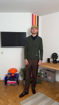 Tumši zaļš džemperis ar ribotu tekstūru, pogu aizdari un priekšējām kabatām, apvienots ar brūniem biksēm un spīdīgām kurpēm. Neitrāls fons.