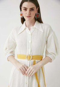 Weißes strukturiertes Kleid mit kragenförmigem Ausschnitt, Vorderknöpfen und einem gelben Gürtel mit einer runden, geflochtenen Schnalle.