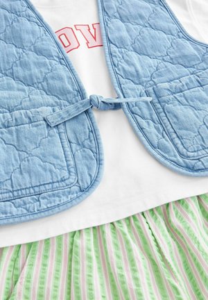 Svijetloplavi quilted prsluk s vezanjem, s dva prednja džepa, složen preko bijele košulje i suknje s zelenim prugama.