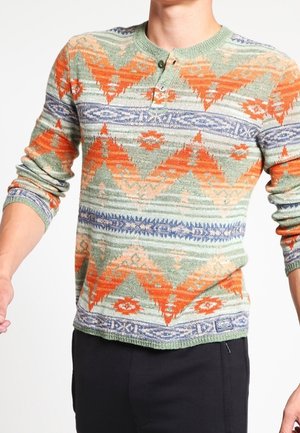 Mand iført langærmet striktrøje med geometriske orange, grønne og blå mønstre samt sorte bukser.