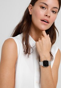 Montre numérique argentée avec un cadran rectangulaire et un bracelet métallique. Portée sur une main avec une chemise blanche à col clair. Design simple et moderne.