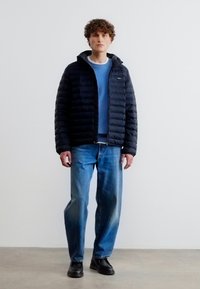 Νεαρός άνδρας που φοράει μαύρο μπουφάν με ραφές πάνω από μπλε πουλόβερ, φαρδιά μπλε τζιν και μαύρα παπούτσια, στέκεται μπροστά σε ένα απλό λευκό τοίχο.