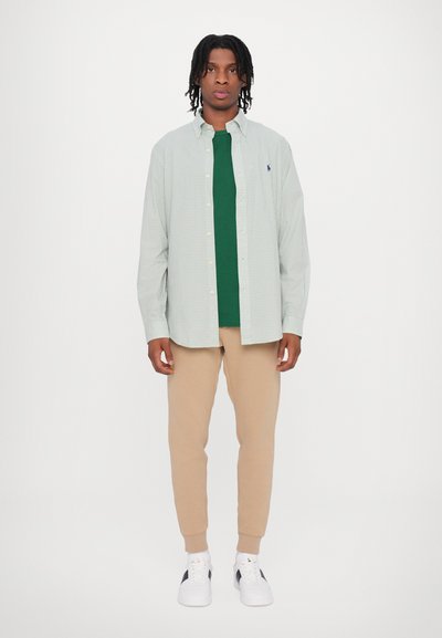 Cămașă cu carouri verzi și albe peste un tricou verde solid, asociată cu pantaloni de trening bej și adidași albi.