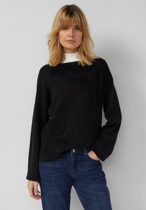 Maglione nero lavorato a maglia con dettaglio floreale strutturato sulla parte frontale, con maniche lunghe, scollo ampio, abbinato a jeans blu scuro.