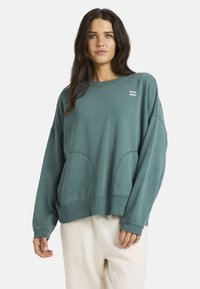 Sudadera de gran tamaño en color verde azulado, con cuello redondo, mangas largas y bolsillos frontales. Presenta un sutil detalle del logo y puños acanalados.