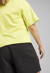 Person som bär en kortärmad gul skjorta och svarta shorts, som visar ryggen och höger arm mot en enkel grå bakgrund.