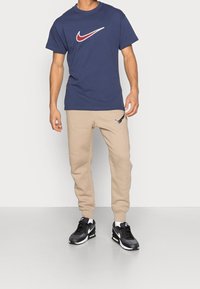 Námořnická modrá triko s kulatým výstřihem a červenobílým logem Nike, v kombinaci s béžovými tepláky se malým stříbrným logem Nike, nošeno s černými teniskami.