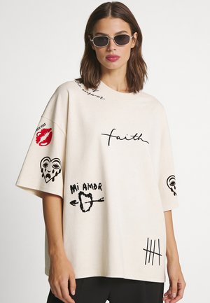 Frau trägt übergroßes weißes T-Shirt mit schwarzen und roten Prints, darunter ein weinendes Herz und Strichlisten, sowie schwarze Sonnenbrille.