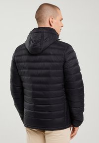 Hombre de perfil con una chaqueta acolchada negra con capucha y pantalones beige, contra un fondo claro y liso.