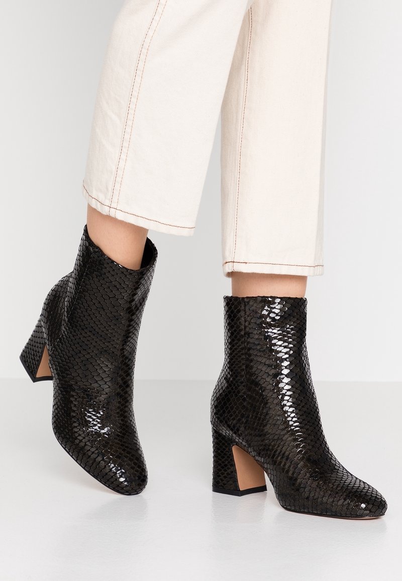Bottes cheville brillantes à motif serpent noir avec talons blocs portées avec un pantalon court écru sur un sol blanc.