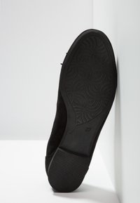 Semelle de chaussure noire inclinée contre un mur blanc, montrant un motif de grip texturé et la taille 37 marquée.