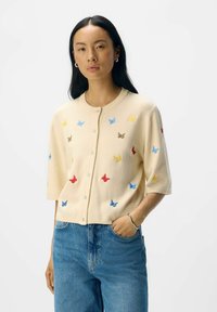 Gilet crème à manches courtes, décoré de broderies de papillons multicolores en bleu, rouge, jaune et marron. Texture légère, avec un devant boutonnière.