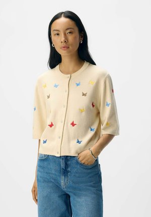 Gilet crème à manches courtes, décoré de broderies de papillons multicolores en bleu, rouge, jaune et marron. Texture légère, avec un devant boutonnière.