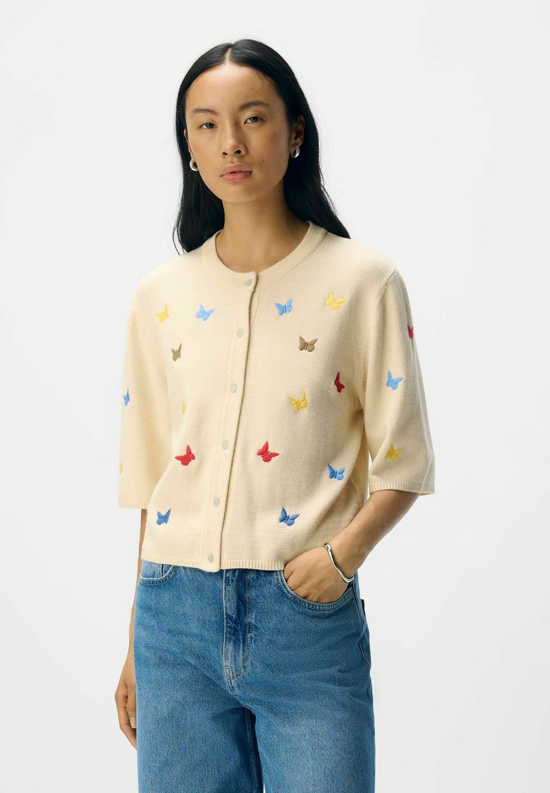 Gilet crème à manches courtes, décoré de broderies de papillons multicolores en bleu, rouge, jaune et marron. Texture légère, avec un devant boutonnière.