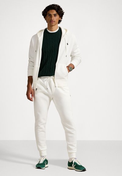 Un sweat à capuche zippé blanc sur un pull en tricot côtelé vert foncé, associé à des joggers blancs et des chaussures de sport vertes. Présence d'un logo sur le sweat.