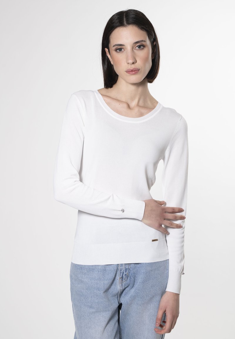 Maglione bianco a maniche lunghe in maglia con scollo rotondo, orlo a coste e dettagli a bottoni sulle maniche; abbinato a jeans in denim azzurro chiaro.