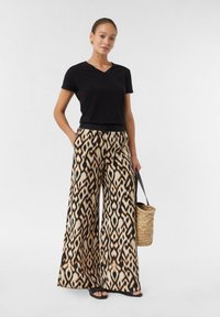Zwarte V-hals T-shirt gecombineerd met rechte broek met een abstract beige en zwart patroon. Model draagt een geweven mandtas.