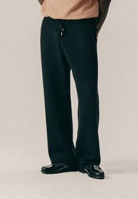 Άτομο που φοράει μαύρο παντελόνι με φαρδιά πόδια και κορδόνι, καθώς και μαύρες δερμάτινες λόφερ, στέκεται μπροστά σε ανοιχτό φόντο.