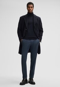Zwarte wollen jas, donkere coltrui, navy textuurbroek en zwarte veterschoenen. Rechte snit, minimale hardware en geen zichtbare patronen.