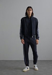 Ung mann som står iført svart hettejakke med glidelås og matchende svarte joggebukser med sidestriper, hvite joggesko, foran en ensfarget grå vegg.