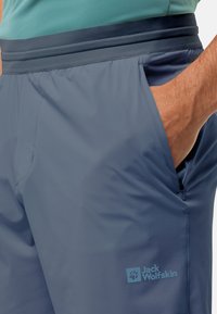 Mann trägt blaue Outdoor-Hosen mit elastischem Bund und hat die Hand in der seitlichen Reißverschlusstasche, das Jack Wolfskin-Logo ist am Oberschenkel sichtbar.