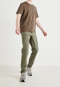 T-shirt in cotone marrone con scollatura rotonda e una piccola grafica sul petto, abbinato a pantaloni dritti verdi e sneaker grigie.