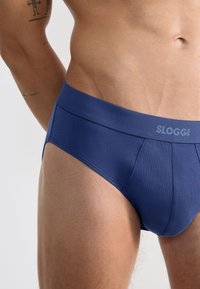 Männlicher Oberkörper in blauen Sloggi-Unterhosen, zeigt einen Teil des Arms mit einem Teil-Tattoo vor einem neutralen Hintergrund.
