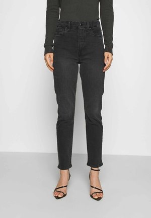 Personne portant un jean skinny noir taille haute, un haut à manches longues foncé et des sandales à talons noires à lanières, debout devant un fond uni.