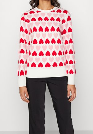 Woman wearing a white sweater with red and pink heart patterns and black pants, standing against a plain light background.