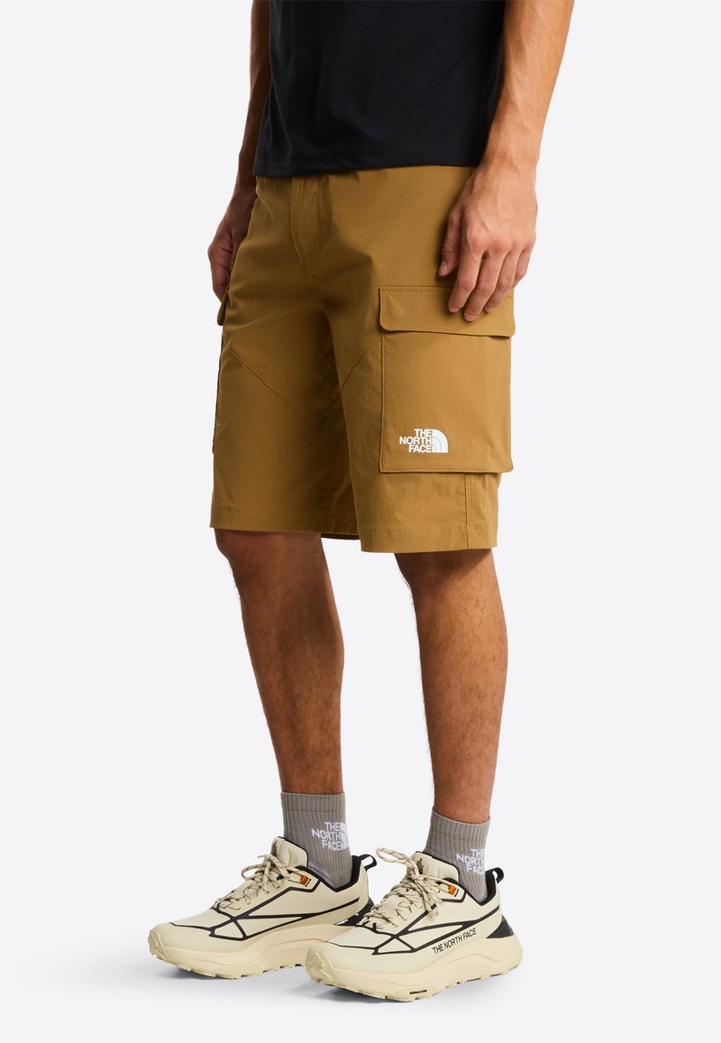 Krótkie spodenki typu cargo w kolorze khaki z wieloma kieszeniami i przeszyciami, połączone z jasnymi trampkami i szarymi skarpetkami. Na spodenkach i butach widoczne logo.