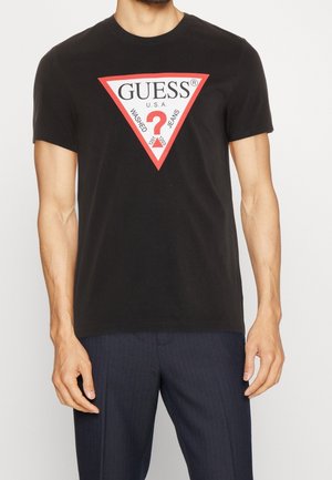 Mand iført sort T-shirt med rød-hvid trekantet Guess-logo og mørke, stribede bukser mod ensfarvet baggrund.