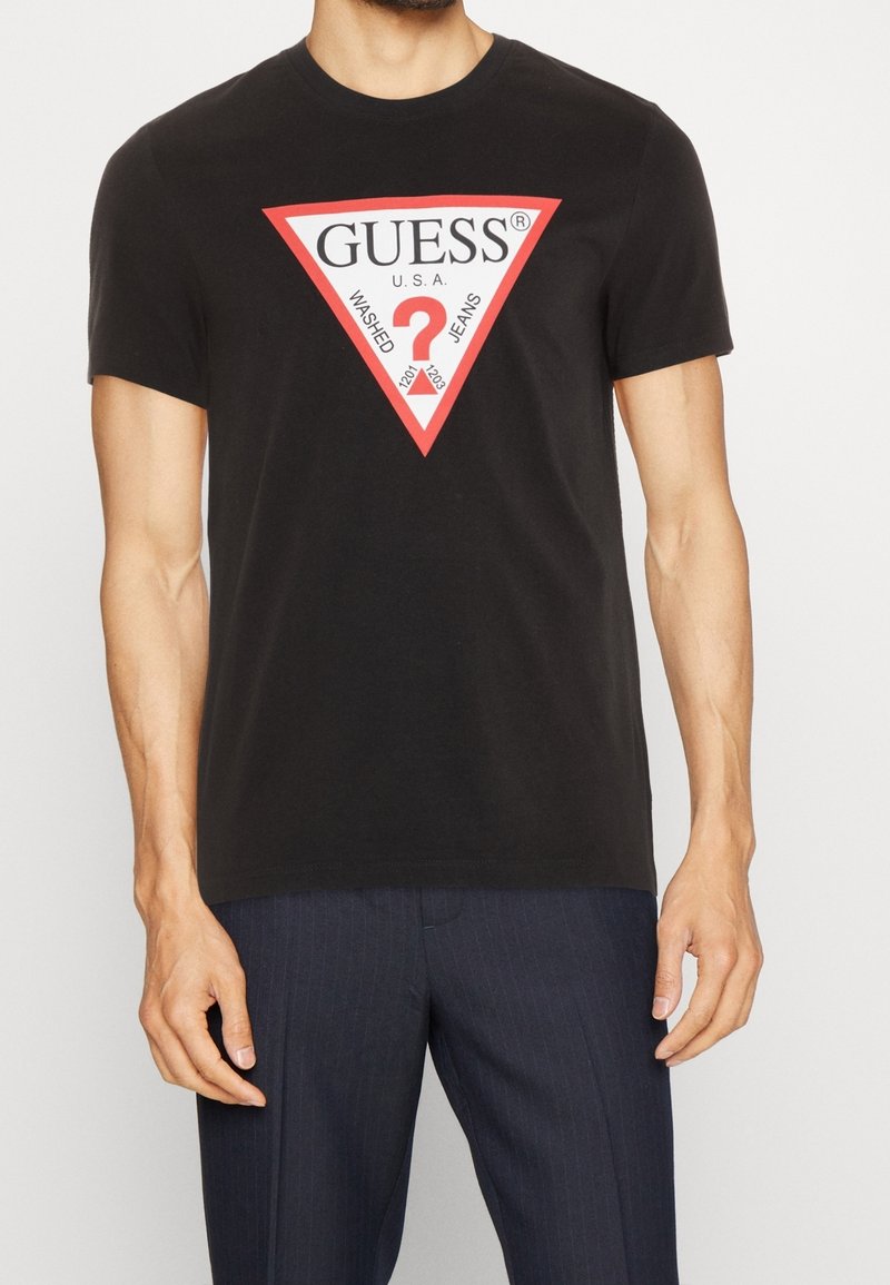 Homme portant un T-shirt noir avec un logo triangulaire Guess rouge et blanc et un pantalon à fines rayures foncé sur un fond uni.