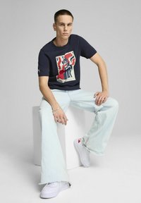 Granatowy T-shirt z graficznym nadrukiem, luźny krój, w połączeniu z jasnoniebieskimi spodniami w szerokim kroju i białymi trampkami z czerwonymi akcentami. Proste tło.
