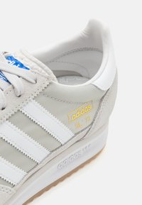 Buty Adidas SL 72 charakteryzują się jasnoszarym materiałem górnym, białymi skórzanymi paskami, beżowym zamszowym obcasem oraz gumową podeszwą.