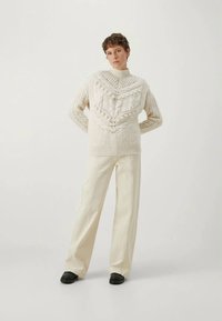 Κρεμ πουλόβερ πλεκτό με υφασμάτινα σχέδια και ψηλό γιακά, σε συνδυασμό με ανοιχτό μπεζ παντελόνι φάρδυ και μαύρα παπούτσια.