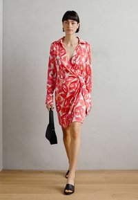 Robe portefeuille dans un motif abstrait rouge vif et rose. Fabriquée en tissu léger, elle présente des manches longues et une taille à nouer. Chaussures noires.