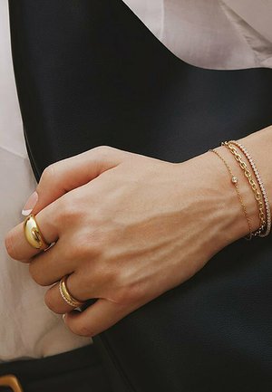 Guld- og sølvarmbånd samt ringe på en hånd, med en struktureret sort læderbaggrund. Ringene har glatte og udsmykkede designs.