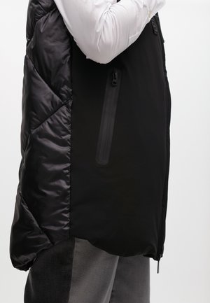 Gilet imbottito nero con texture lucida sul lato sinistro e finitura opaca sul lato destro, dotato di tasca con zip e design orlo asimmetrico.