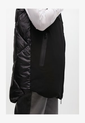 Gilet imbottito nero con texture lucida sul lato sinistro e finitura opaca sul lato destro, dotato di tasca con zip e design orlo asimmetrico.