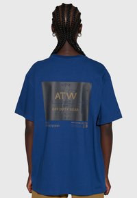 Blaues Baumwoll-T-Shirt mit weiter Passform, auf der Rückseite mit einem schwarzen Grafikdruck versehen. Der Text beinhaltet "ATW" und "OFF DUTY GEAR" in Gold.