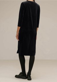 Schwarzes Samtkleid mit Dreiviertelärmeln und geradem Schnitt, getragen mit schwarzen Strumpfhosen und Schuhen. Glatte Textur und schlichtes Design.